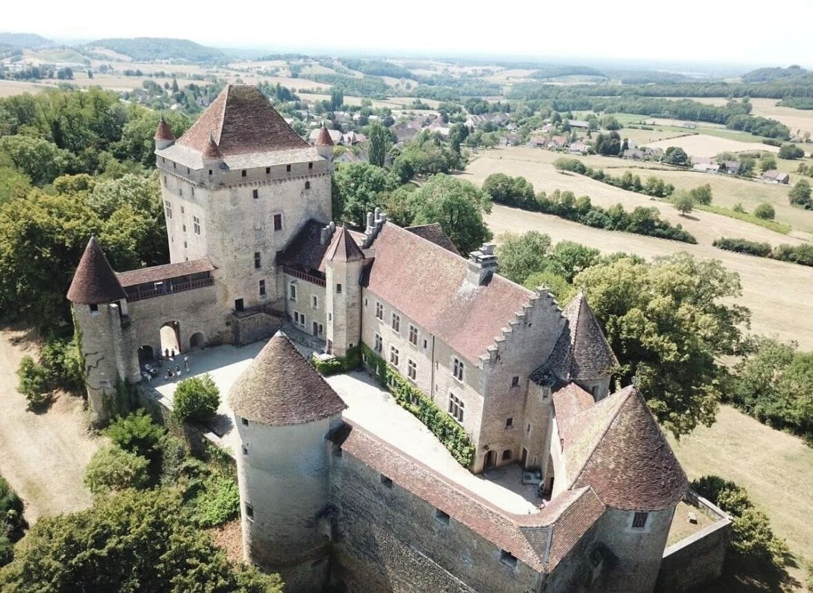 Un château doté de nombreuses tours et d’une cour, entouré de verdure et de champs.