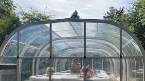 Ein von Glas umschlossener Whirlpoolbereich mit gewölbtem Dach, umgeben von Bäumen.