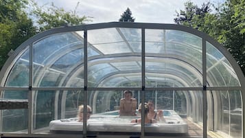 Un espace abrité avec un jacuzzi, entièrement vitré et doté d’un toit incurvé, entouré d’arbres.
