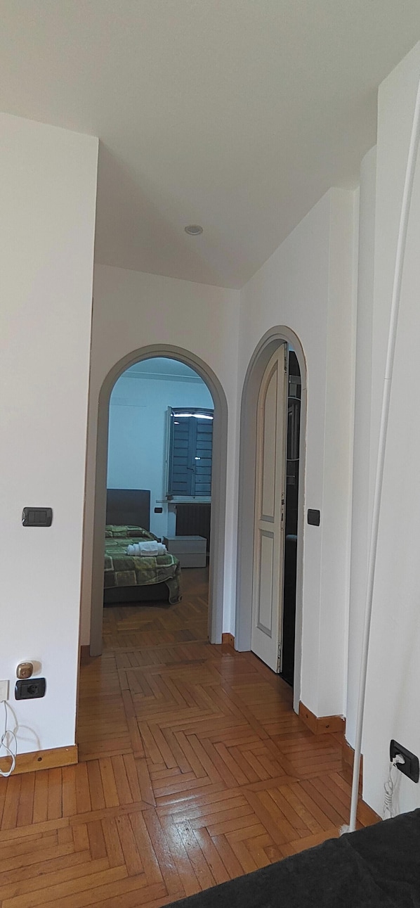 Un couloir dotĂ© dâun plancher en bois, de murs blancs et de deux portes cintrĂ©es donnant sur une chambre Ă coucher Ă©quipĂ©e dâun lit et dâun radiateur.