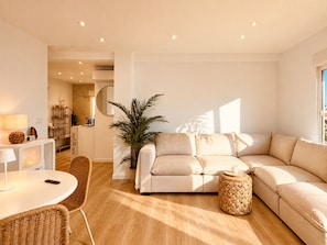 A modern living room with a beige sofa, a round dining table, wicker chairs, a potted plant, and a kitchen area in the background. - La Goleta Apartment 5mins walk from beach (Dénia)