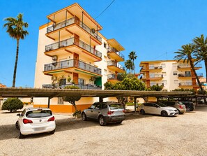 A multi-story residential building with balconies, a parking area with several cars, and palm trees. - La Goleta Apartment 5mins walk from beach (Dénia)