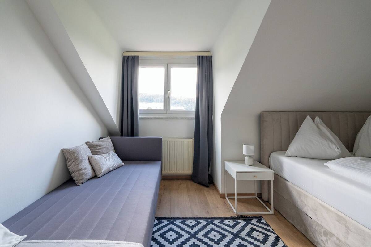 A modern, compact room with a bed, a sofa, a small bedside table, and a window with curtains.