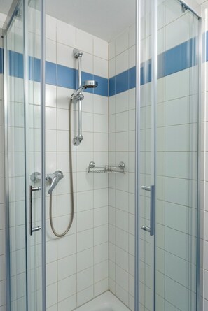 A glass-enclosed shower stall with white and blue tiles, a handheld showerhead, and a wall-mounted faucet. - FarawayHomes Studio Leoben K105 (Donawitz)
