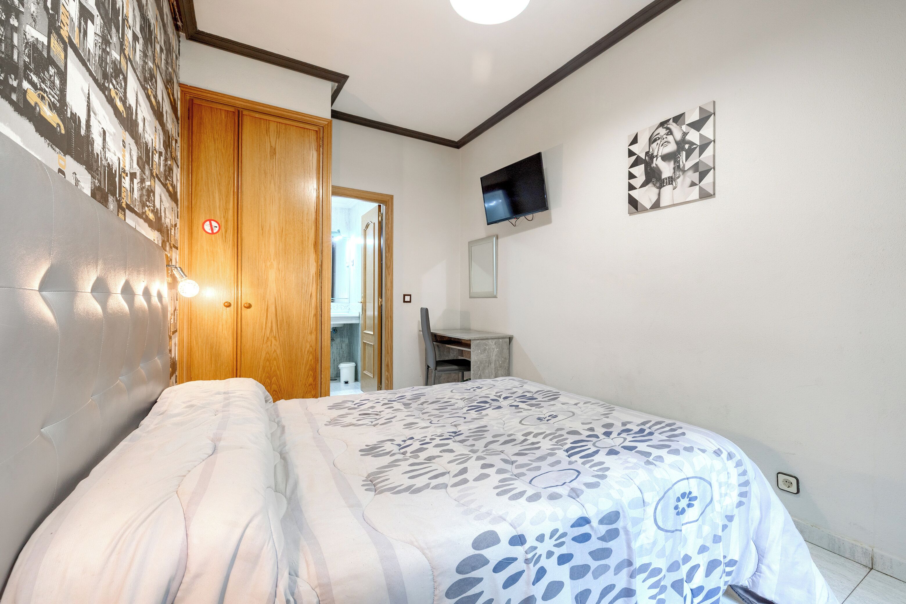 Un dormitorio con cama, una puerta de madera, un escritorio, una silla, un televisor y una pared con un mural de paisaje urbano.