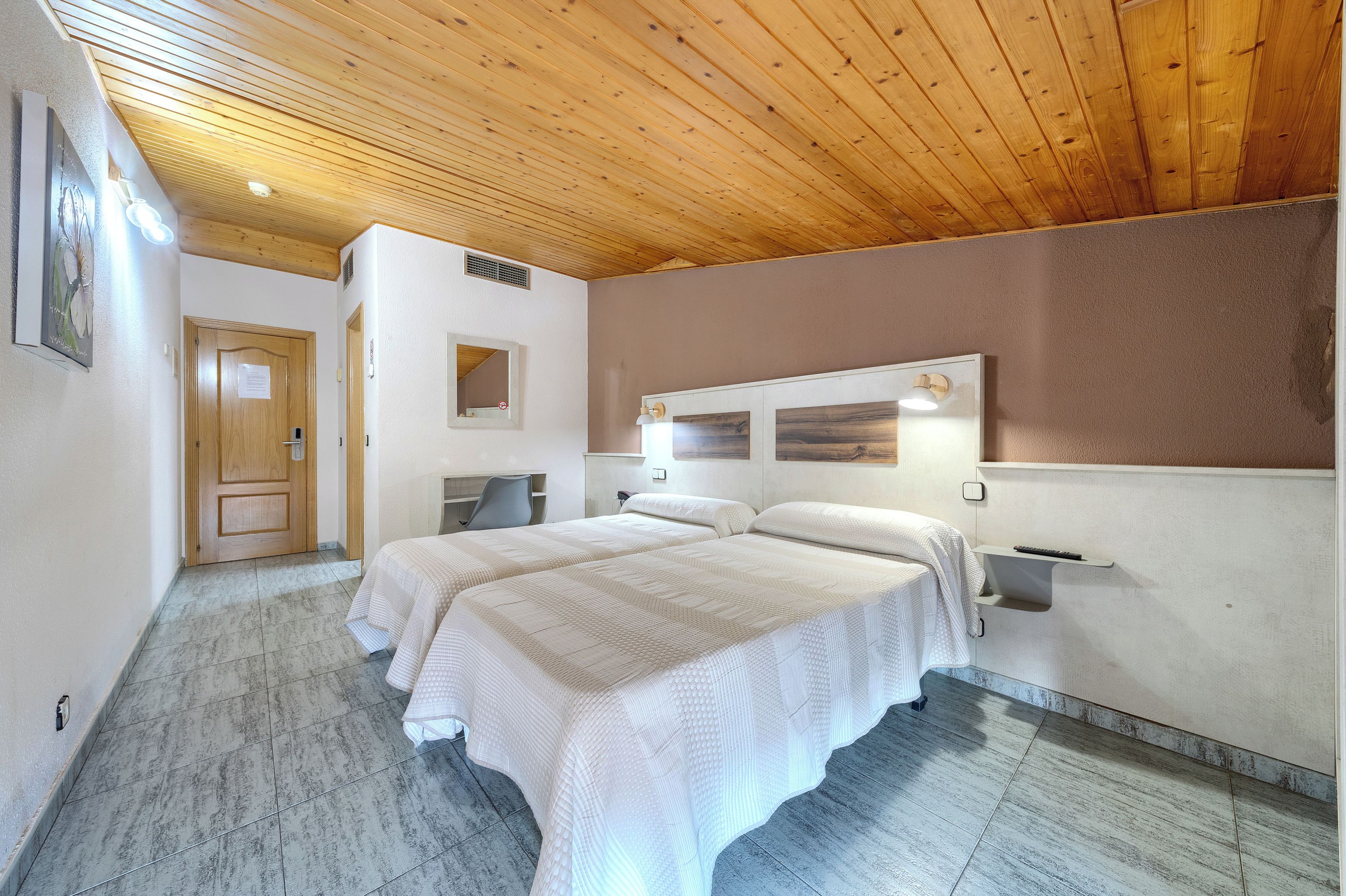A hotel room with a wooden ceiling, a bed with white bedding, a desk, and a chair.