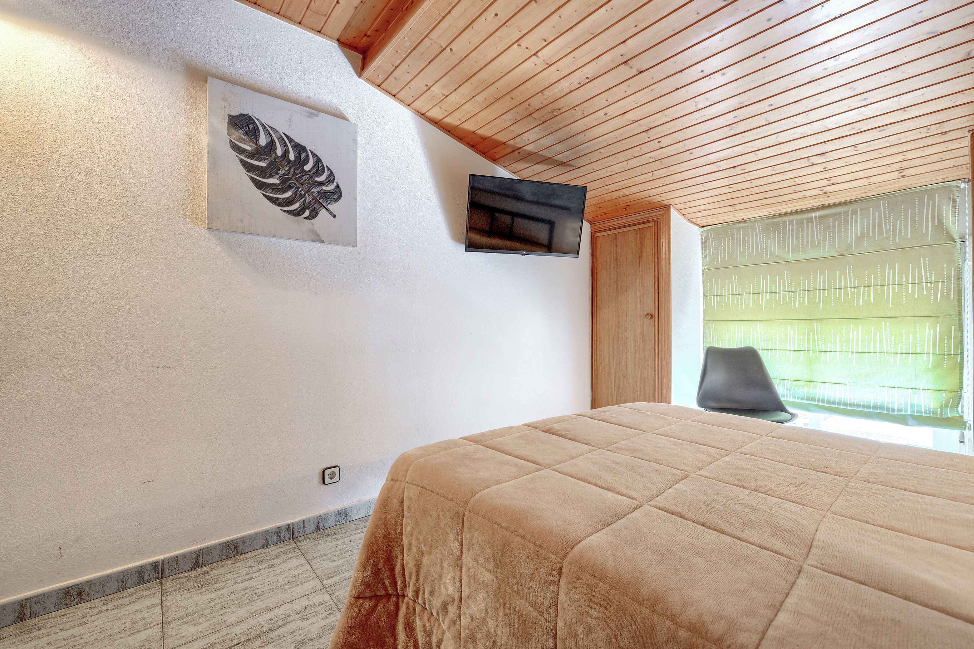 Quarto com cama, televisão na parede, uma obra de arte emoldurada e janela com persianas.