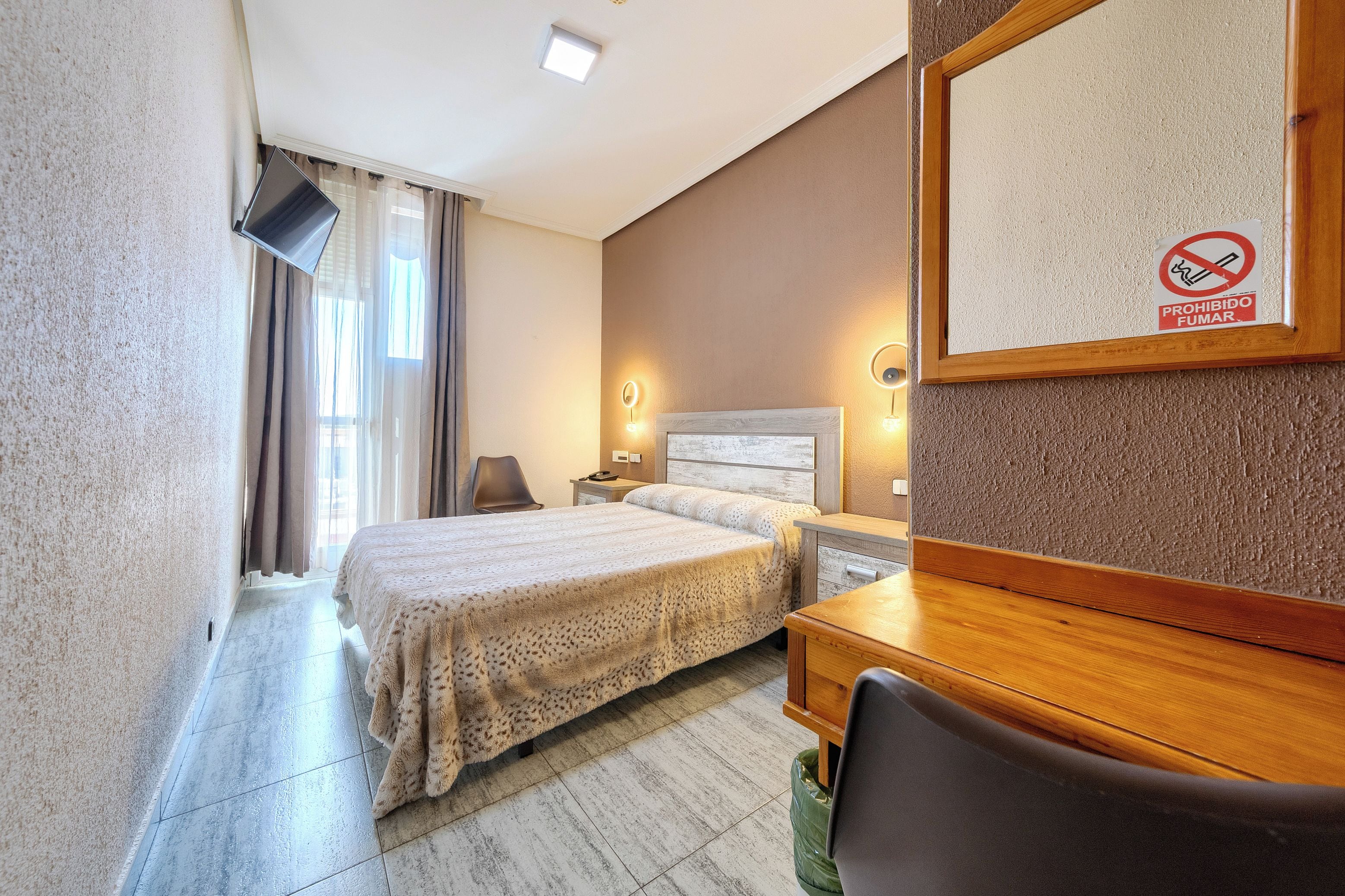 A hotel room with a bed, a desk, a chair, a TV mounted on the wall, and a no-smoking sign.