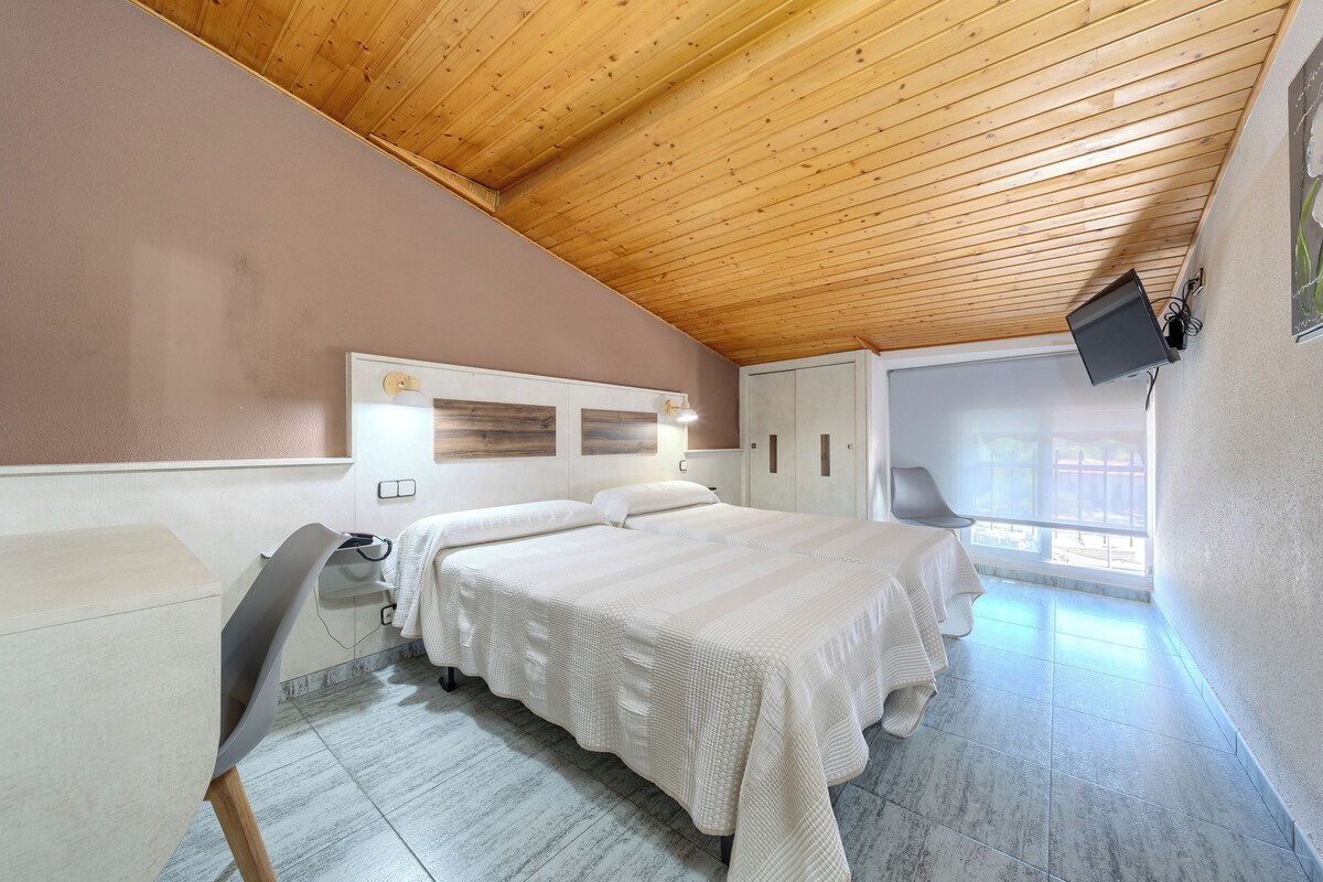 A bedroom with a wooden ceiling, a bed with white bedding, a desk, a chair, a TV mounted on the wall, and a window with a view.
