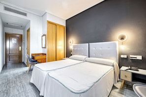 A hotel room with a large bed, a desk with a chair, a wardrobe, and a bedside table with a phone.