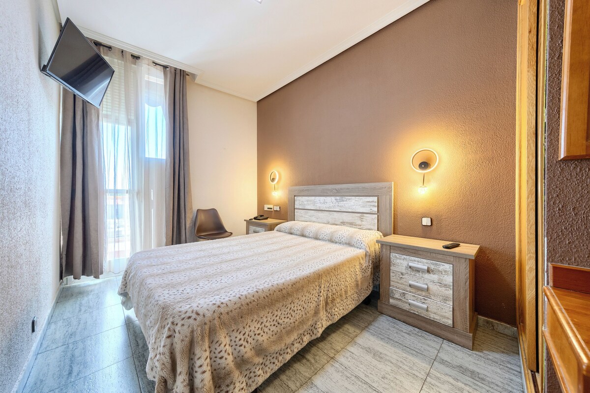 A hotel room with a bed, bedside tables, a flat-screen TV mounted on the wall, and a window with curtains.