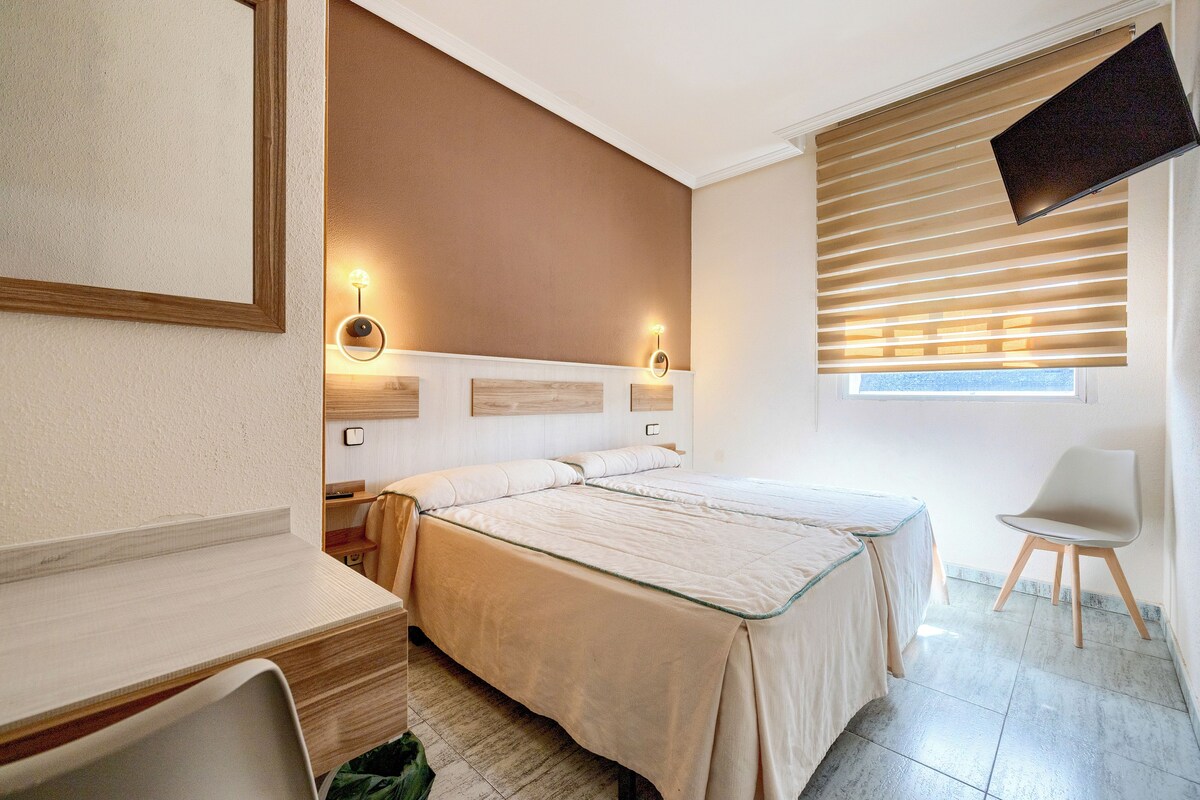 A hotel room with a bed, bedside tables, a desk, a chair, and a window with blinds.