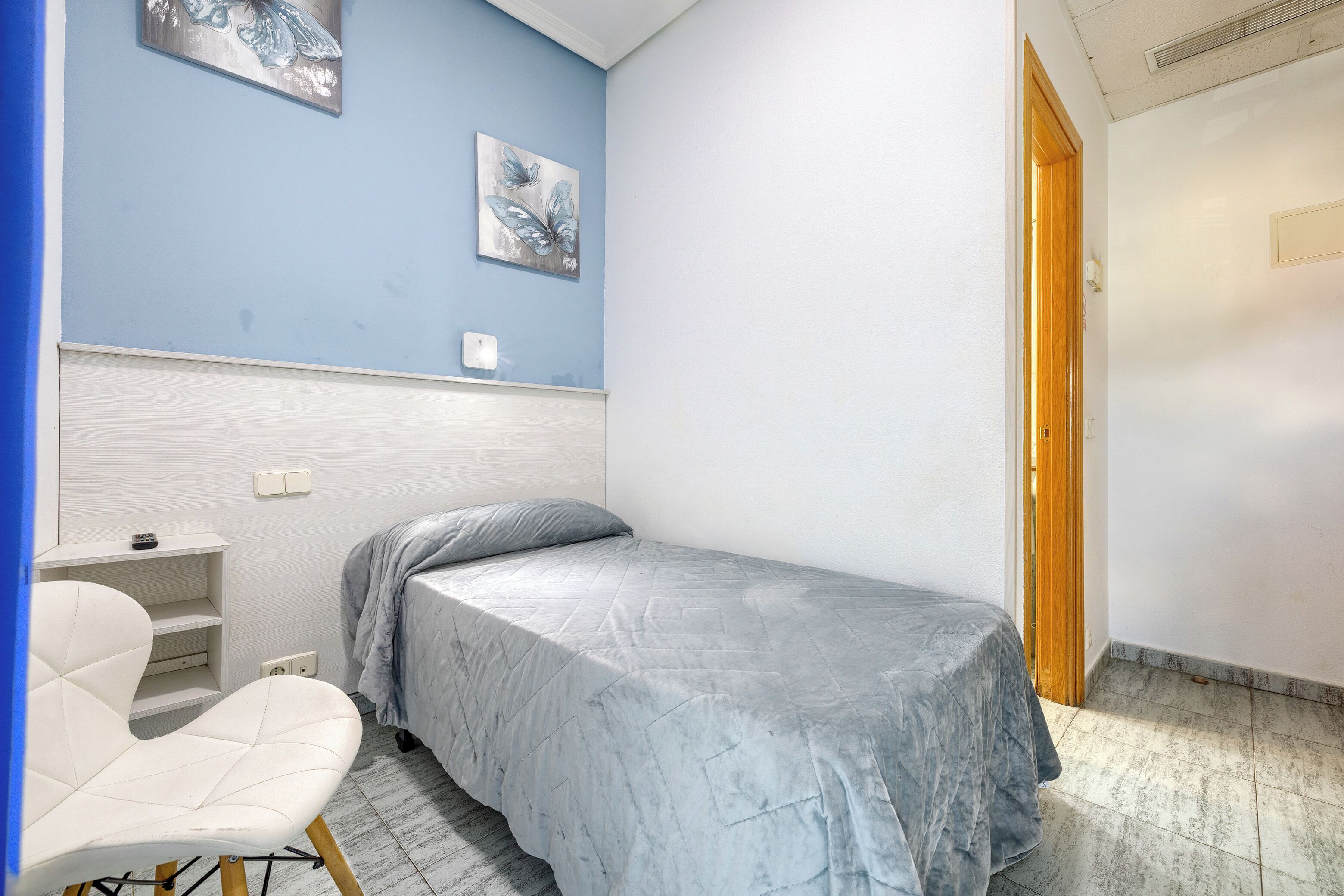 Une petite chambre avec un lit simple, un mur bleu, une chaise blanche et une porte en bois.