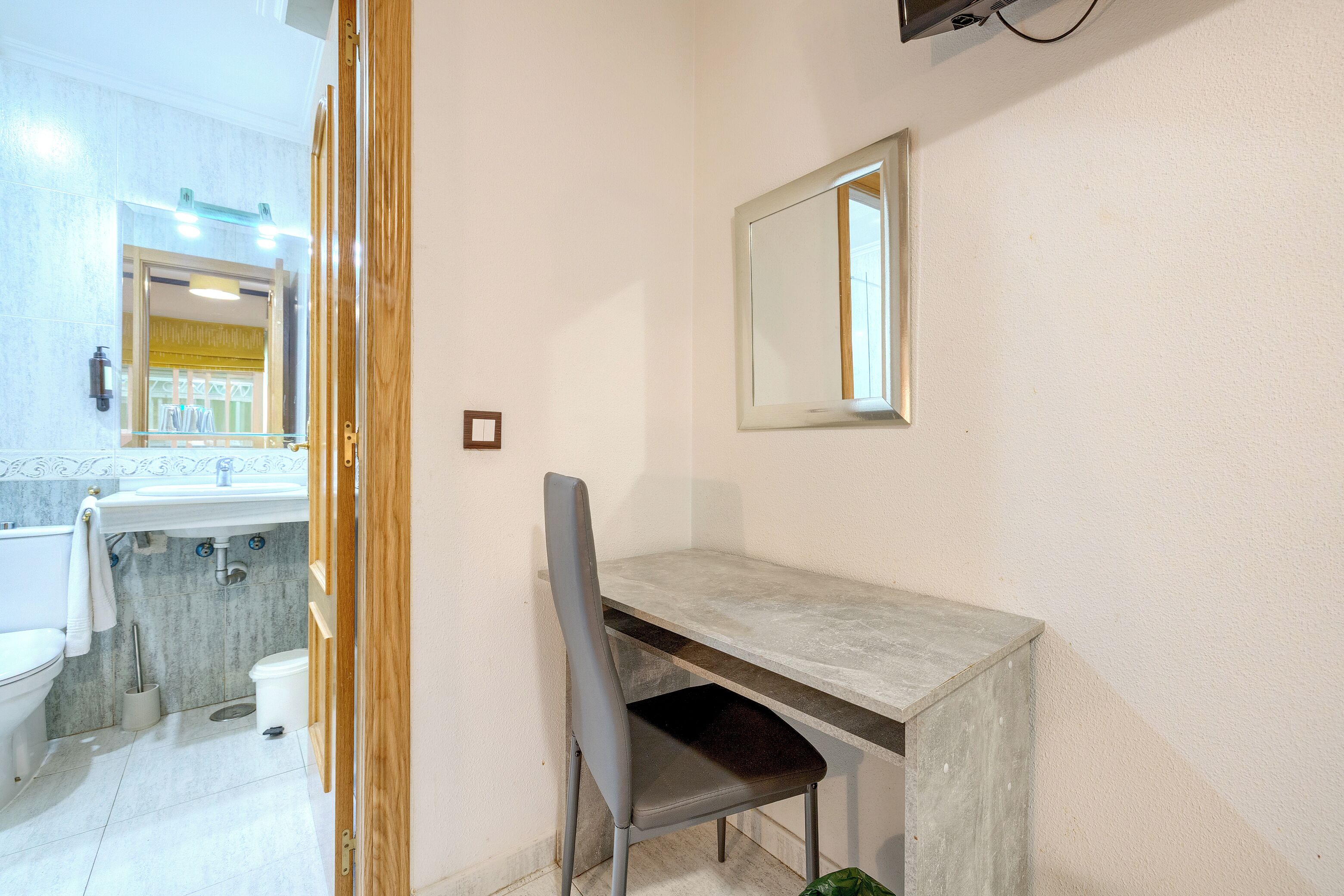 A bathroom with a toilet, sink, mirror, and a desk with a chair.