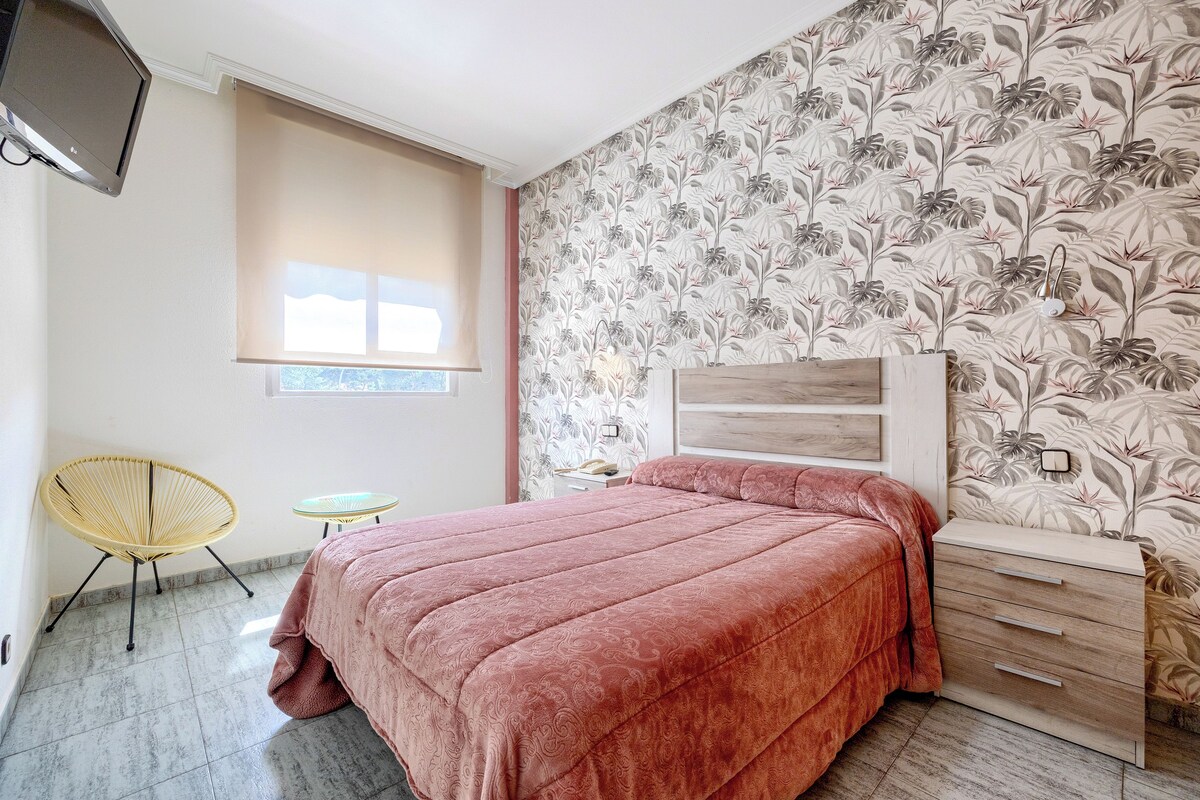 A bedroom with a bed, a bedside table, a chair, a small table, and a TV mounted on the wall.