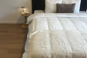 En pænt redt seng med et hvidt dynebetræk og et vævet tæppe, et sengebord med en lampe og en lille stak bøger.
