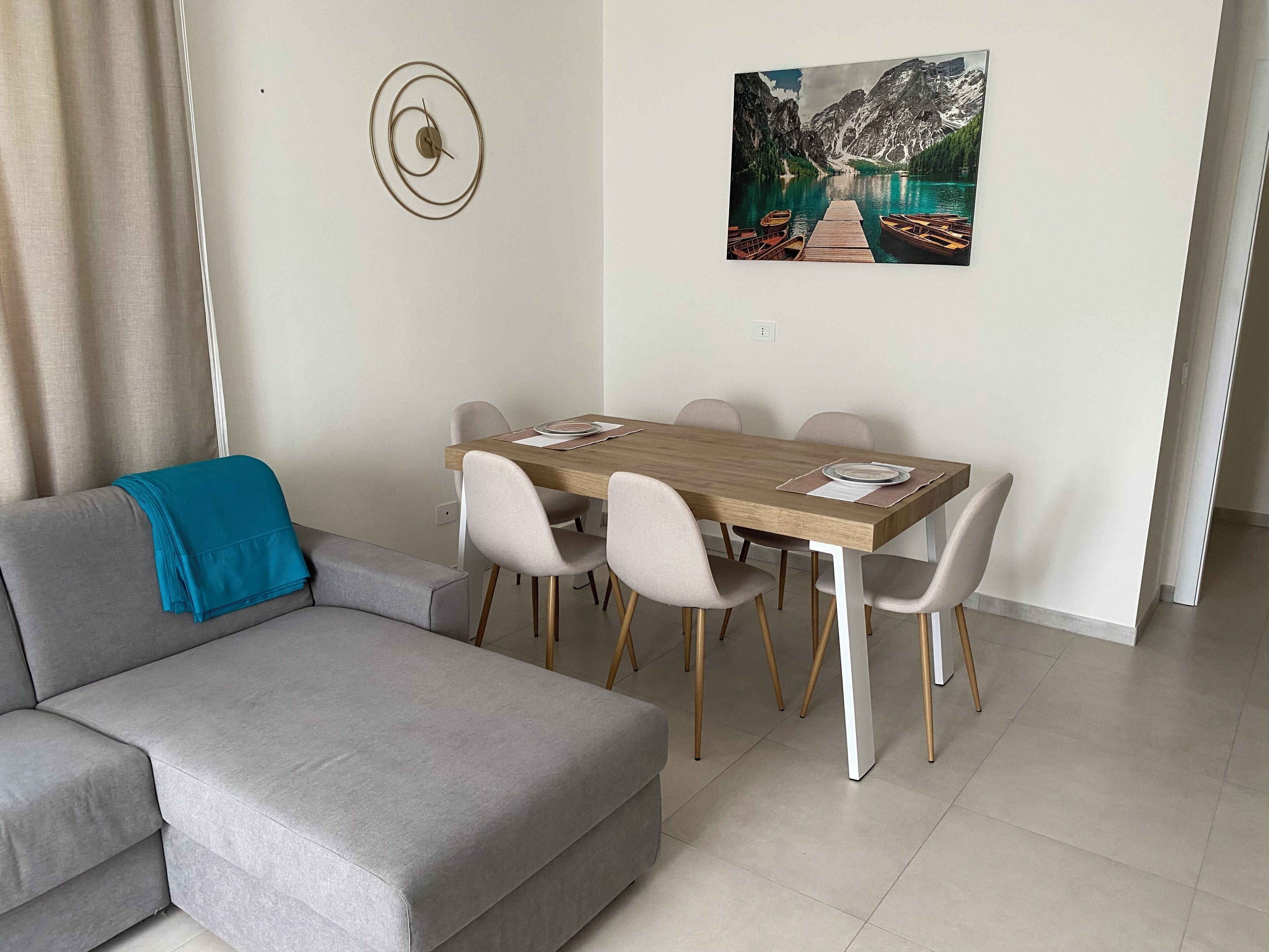 A modern living room with a grey sofa, a wooden dining table with chairs, a painting of a mountain lake, and a wall clock.