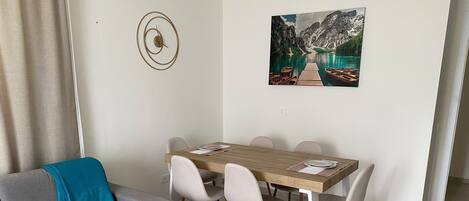 A modern living room with a grey sofa, a wooden dining table with chairs, a painting of a mountain lake, and a wall clock.