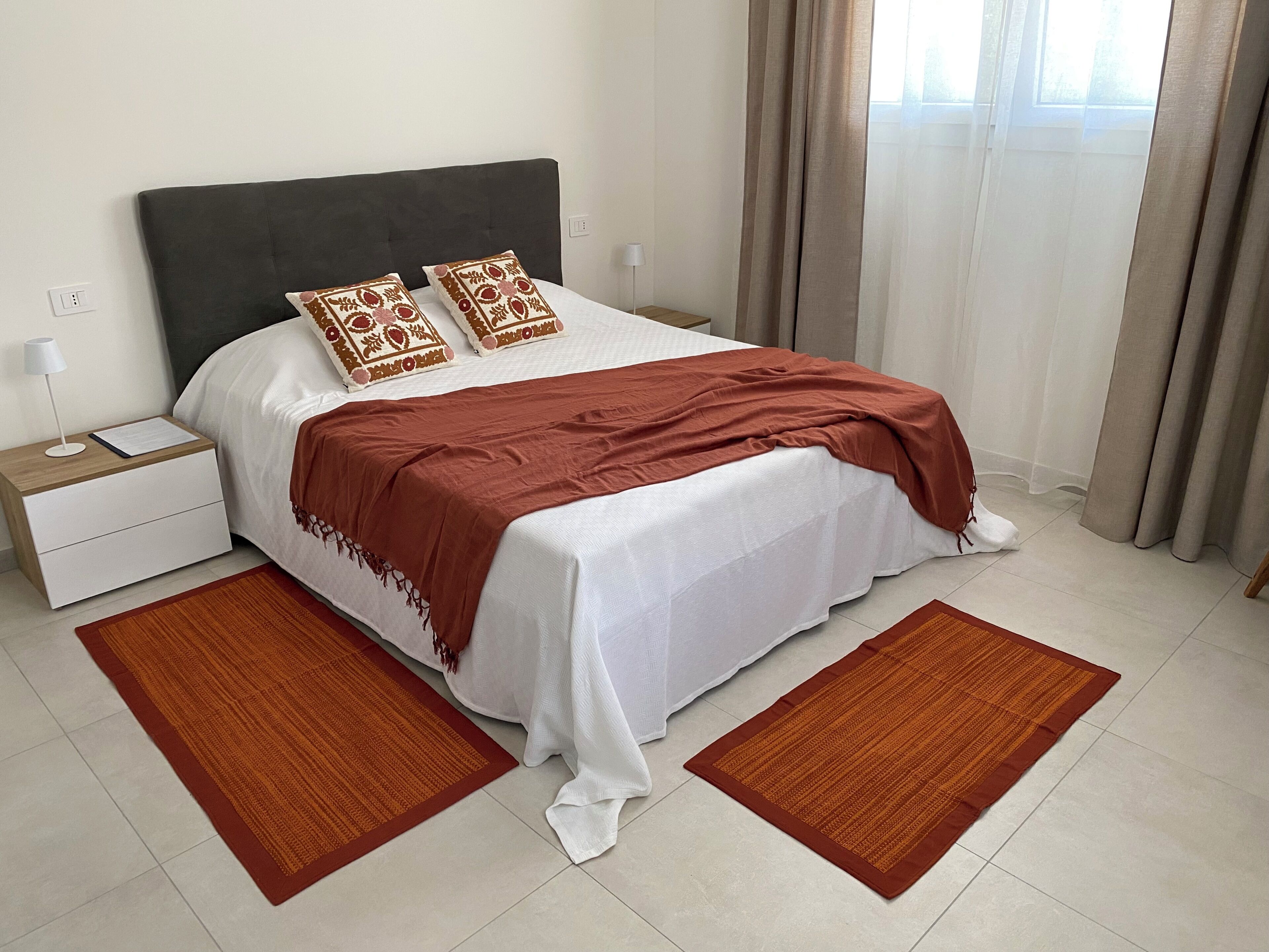 Un lit bien fait, recouvert d’une couette blanche et d’un plaid marron, flanqué de deux tapis marron, d’une table de chevet avec une lampe et d’une fenêtre avec des rideaux.