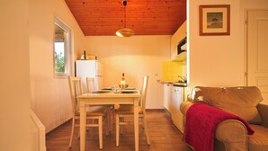 A dining area with a wooden table and chairs, a kitchen with a yellow backsplash, and a brown sofa with a red blanket. - PRIVILEGE 2-Bedroom Cottages (Parentis-en-Born)