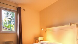 A bedroom with a bed, a window with curtains, a bedside table, and a wall-mounted light. - PRIVILEGE 2-Bedroom Cottages (Parentis-en-Born)