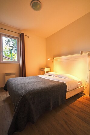 A bedroom with a bed, a window with curtains, a bedside table, and a wall-mounted light. - PRIVILEGE 2-Bedroom Cottages (Parentis-en-Born)