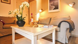 A living room with a white coffee table, a sofa, a potted orchid, a bottle of wine, and a framed picture on the wall. - PRIVILEGE 2-Bedroom Cottages (Parentis-en-Born)