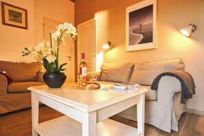 A living room with a white coffee table, a sofa, a potted orchid, a bottle of wine, and a framed picture on the wall. - PRIVILEGE 2-Bedroom Cottages (Parentis-en-Born)