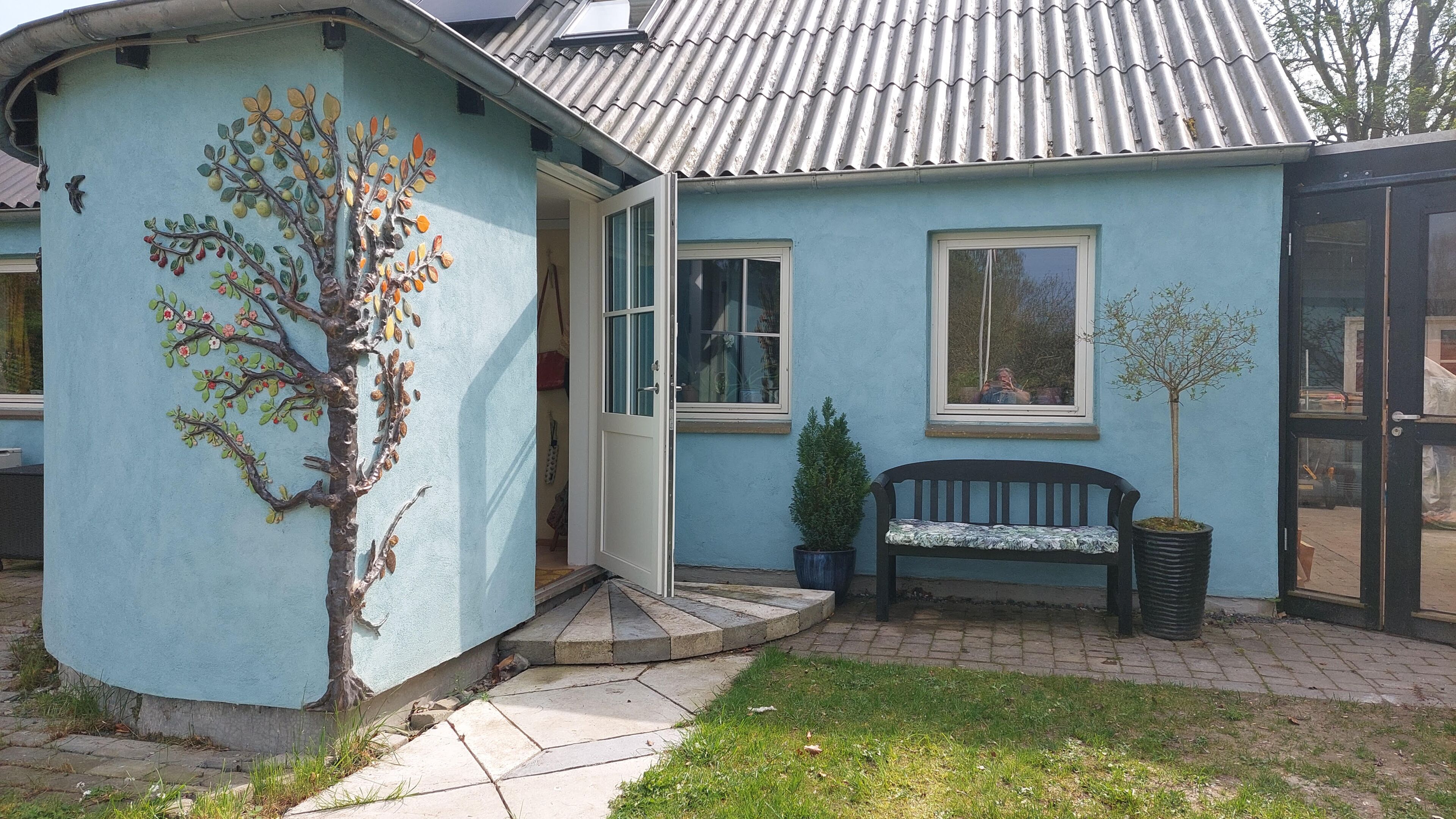 Een huis met een blauwe gevel, een muurschildering van een boom, een bank en een bloempot.