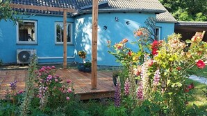 Ein blaues Haus mit einer Holzterrasse, einem Garten voller Blumen und einem Vogelgemälde an der Wand.
