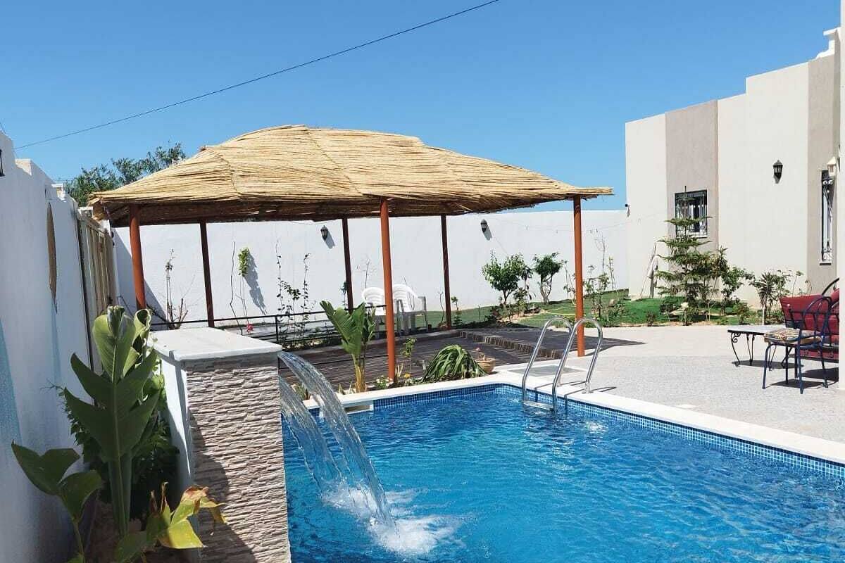 A swimming pool with a water feature, a thatched roof structure, and surrounding greenery.