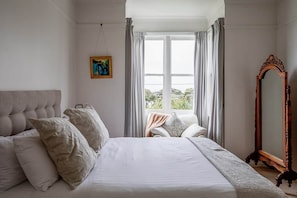 Ein Schlafzimmer mit einem großen Bett, einem Sessel, einem Spiegel und einem Fenster mit Vorhängen.