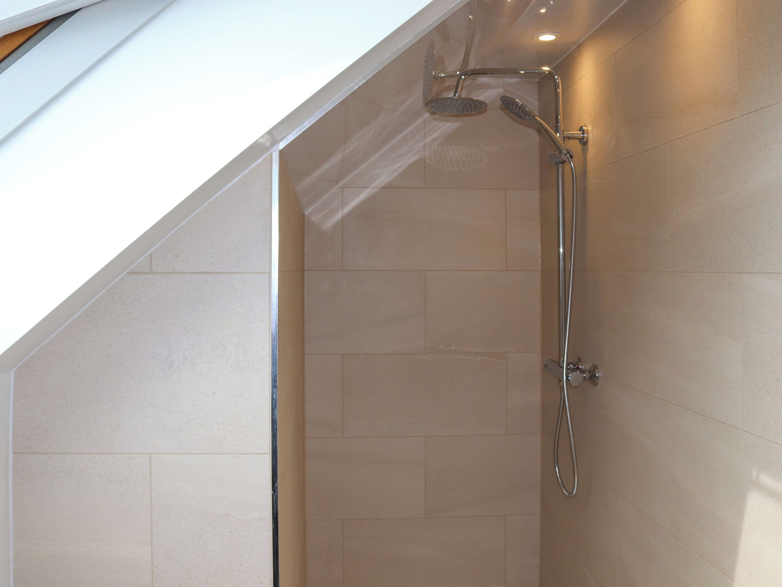 A shower with a handheld showerhead and a wall-mounted faucet in a room with tiled walls and a sloped ceiling.