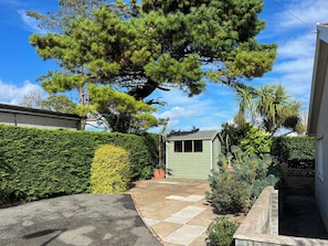 A residential property with a paved driveway, a green shed, and a variety of plants including a large pine tree and a neatly trimmed hedge. - 3 bedroom accommodation in Benllech (Benllech)