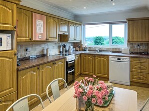 A modern kitchen with wooden cabinets, a built-in oven, a dishwasher, and a window providing a view of greenery. - 3 bedroom accommodation in Benllech (Benllech)