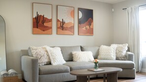 Un salón con un sofá gris, almohadones blancos y esponjosos, una mesa de centro redonda y tres obras de arte con motivos desérticos.