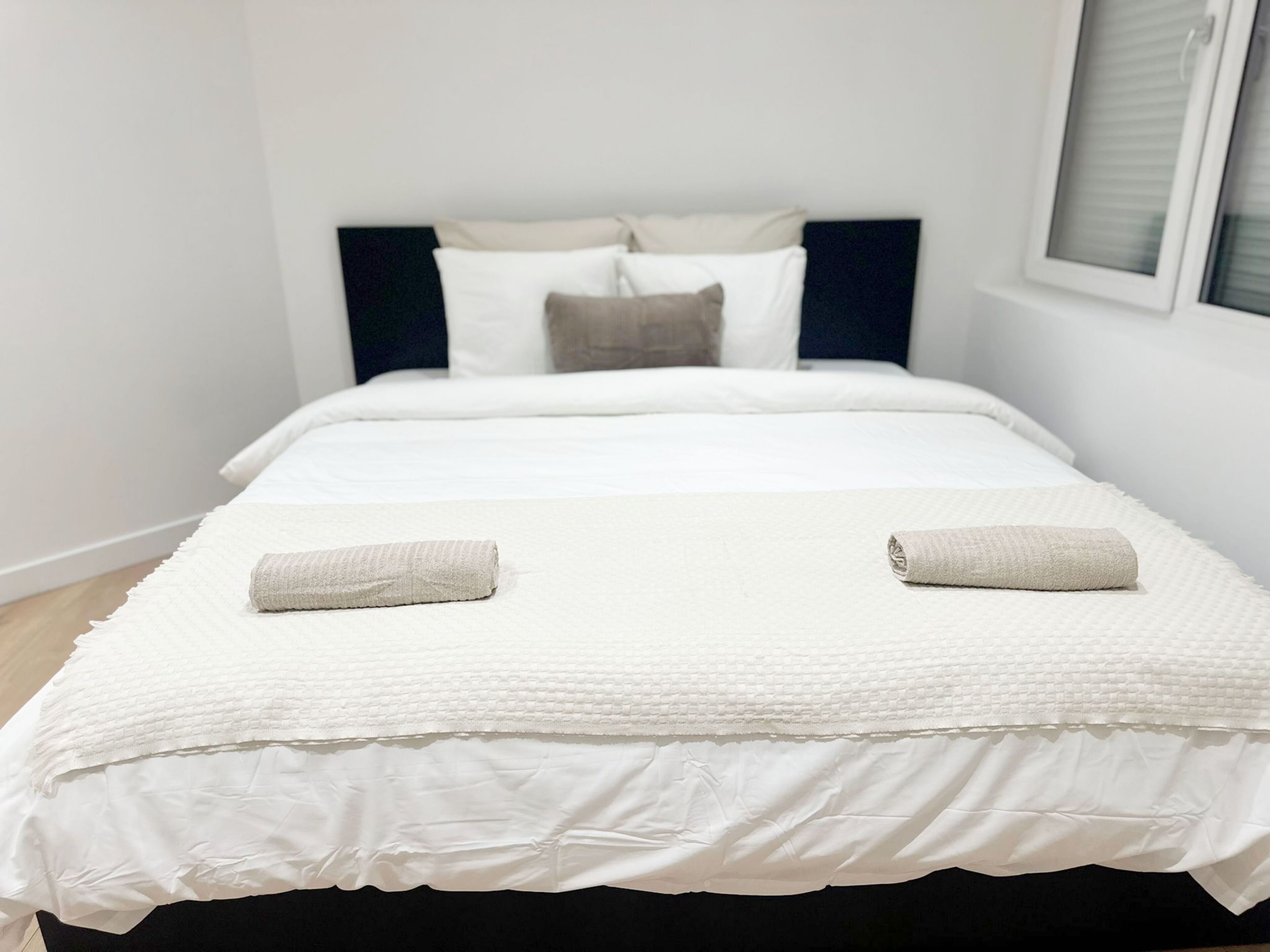 A neatly made bed with a white duvet, two rolled towels, and a dark headboard.