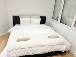 Ein ordentlich bezogenes Bett mit weißer Bettwäsche und zwei aufgerollten Handtüchern, ein dunkles Kopfteil sowie ein Fenster mit Jalousien.