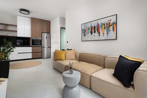 Ein modernes Wohnzimmer mit einem beigefarbenen Sofa, einem runden Couchtisch und einer Küchenzeile mit Holzausbau und einem Kühlschrank.