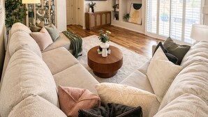 Ett vardagsrum med en stor hörnsoffa, ett soffbord, en tv som är monterad på väggen och ett stort fönster med persienner.