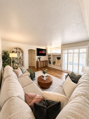 Ein Wohnzimmer mit einem großen Ecksofa, einem Couchtisch, einem an der Wand montierten Fernseher und einem großen Fenster mit Jalousien.