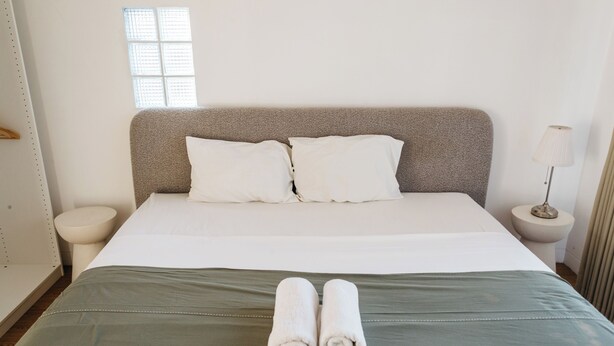 A neatly made bed with pillows, a pair of rolled towels, and a bedside lamp.