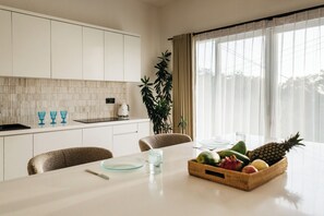 A modern kitchen with white cabinets, a dining table with a fruit basket, and a large window with sheer curtains. - New 2BR Apartment near Dreamland Beach (Kuta)