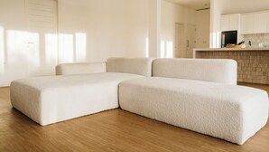A modern living room with a sectional sofa, wooden flooring, and a kitchen area with white cabinets.