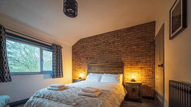Un dormitorio con pared de ladrillo, una cama grande con ropa blanca, dos lámparas de noche y una ventana con cortinas.