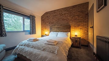 Une chambre avec un mur en briques, un grand lit recouvert de draps blancs, deux lampes de chevet et une fenêtre avec des rideaux.