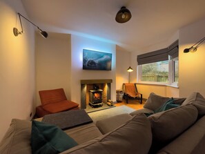A cozy living room with a fireplace, a sofa, a chair, and a TV mounted on the wall. - Lovely mews cottage in the western Lake District (Eskmeals)