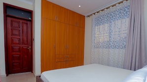 A bedroom with a bed, wooden wardrobe, and patterned curtains. - Imaani Living Buziga (Kampala)