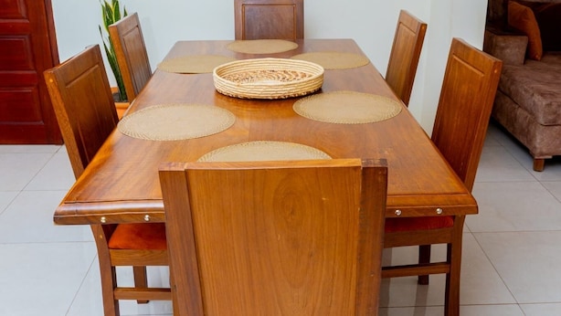 A wooden dining table with six chairs, a woven placemat, and a painting above it. - Imaani Living Buziga (Kampala)