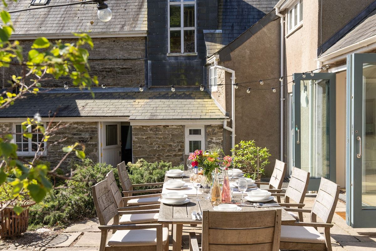 An outdoor dining area with a table set for six, surrounded by stone walls and a glass door leading to a garden.